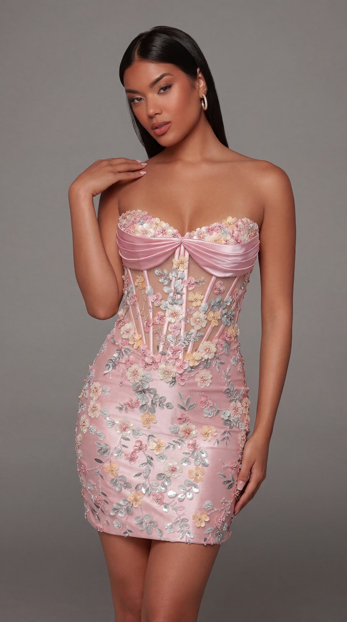 Wearing the Alyce 40139 Short Homecoming Dress in Rosewater, this woman showcases a strapless, fitted mini silhouette adorned with delicate floral appliques and a sheer corset bodice, paired with hoop earrings and posing against a plain gray background.